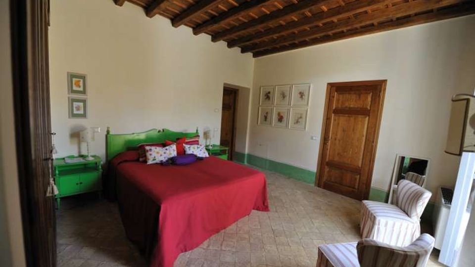 Camera da letto in stile rustico con soffitto in legno a travi a vista, pavimento in piastrelle e pareti bianche con fascia verde. Al centro c’è un letto matrimoniale con copriletto rosso e testiera verde, affiancato da comodini coordinati; sulla destra si vedono due poltroncine a righe e una porta in legno.
