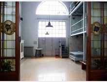 Interno di un ampio spazio tipo loft con grandi finestre ad arco e a griglia sopra una porta centrale, illuminato da due lampade a sospensione. Ai lati ci sono porte in legno con vetri decorati, una scala metallica che porta a un soppalco e arredi essenziali su pavimento chiaro.