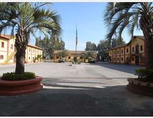 Ampio cortile pavimentato circondato da edifici bassi color ocra con finestre regolari e scale esterne, con palme in grandi fioriere ai lati. Al centro si trova un pennone con bandiera e sullo sfondo si vedono alberi oltre gli edifici.