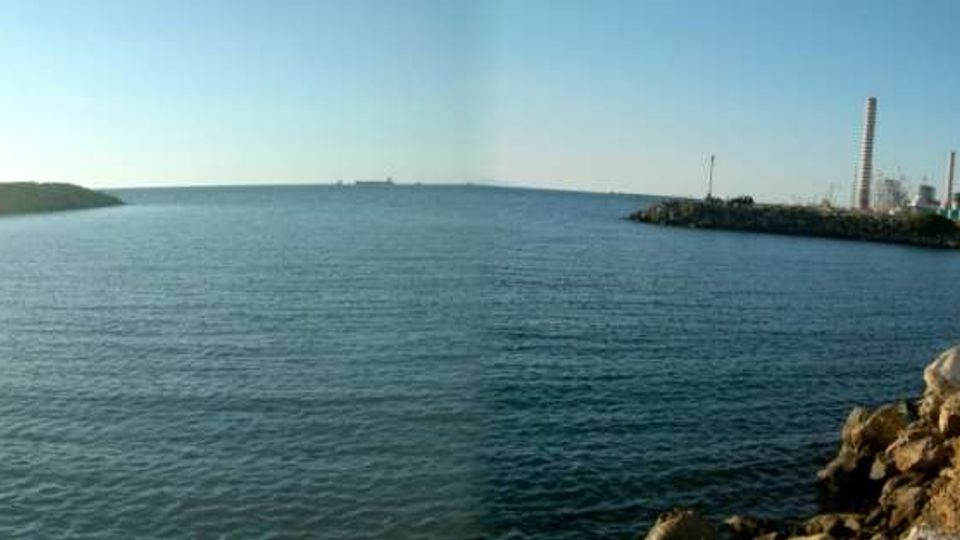 Veduta panoramica di un ampio tratto di mare con costa rocciosa in primo piano e una bassa penisola all’orizzonte. Sul lato destro si vede un’area industriale sul litorale con alti camini e strutture portuali.