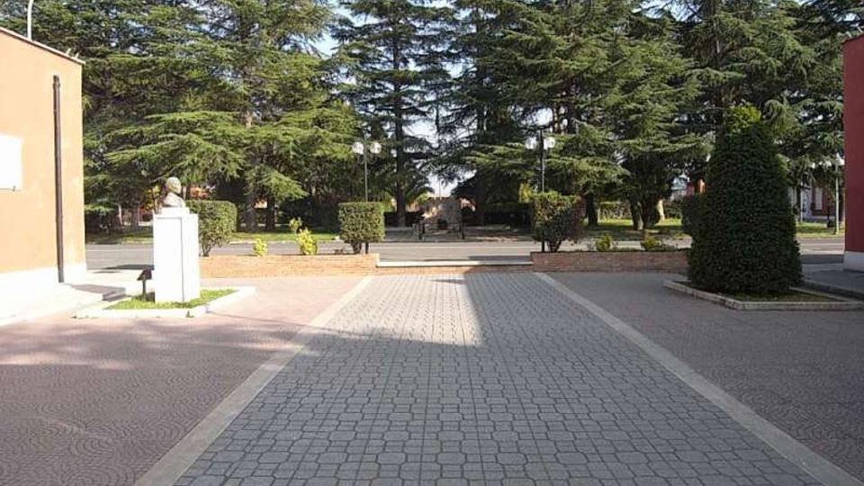 Piazzale pedonale pavimentato con blocchetti e corsie laterali in pietra, affiancato da siepi potate e lampioni. Sullo sfondo si vedono alti alberi sempreverdi e, a lato, edifici con facciate semplici e una piccola statua su piedistallo.