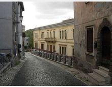 Strada in discesa lastricata in pietra tra edifici storici, con un grande palazzo chiaro a due piani con finestre ad arco sullo sfondo. Ai lati si vedono facciate intonacate, un lampione a muro e una ringhiera lungo il bordo della strada sotto un cielo nuvoloso.
