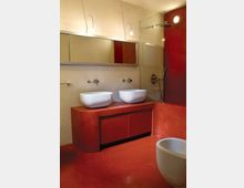 Bagno moderno con pavimento e rivestimenti in resina rosso intenso, mobile lavabo coordinato con due lavabi a bacinella e rubinetteria a parete sotto uno specchio lungo. A destra è visibile una doccia con parete in vetro e soffione, con un sanitario bianco in primo piano.