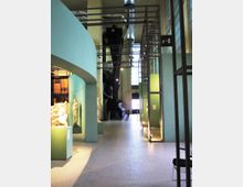 Interno di un museo in un ampio corridoio con pareti verde acqua, pavimento a mosaico chiaro e grandi condotte metalliche a vista sul soffitto. Lungo il percorso si vedono teche e piedistalli con statue, strutture metalliche verticali e un’alta finestra sul fondo che illumina lo spazio.