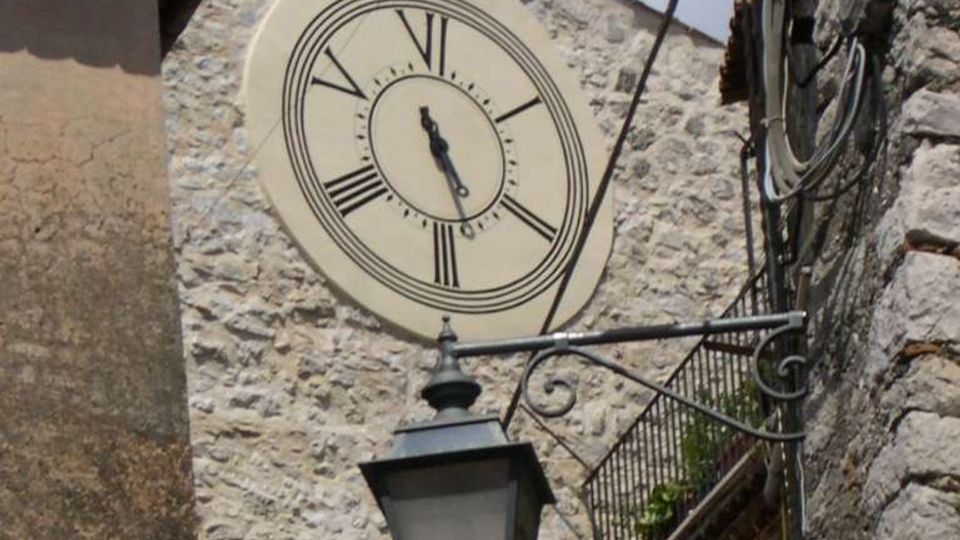 Facciata in pietra di un edificio storico con grande orologio rotondo a numeri romani e una campana in una struttura in ferro sopra il tetto. In primo piano si vede un lampione sospeso e, a destra, un balcone con ringhiera e cavi lungo il muro di una stretta via tra edifici.