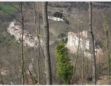 Veduta panoramica di un borgo collinare con case in pietra e tetti in coppi, osservato attraverso tronchi e rami di alberi in primo piano. Sulla destra si distingue un grande edificio storico con mura chiare e tetto rosso, circondato da vegetazione e pendii boscosi.