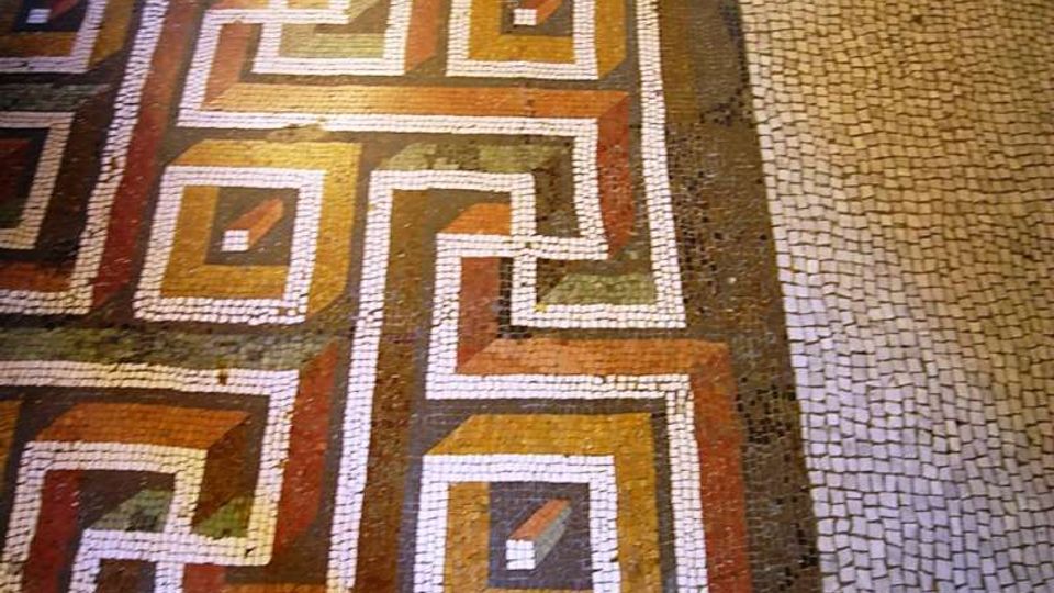 Pavimento a mosaico con motivi geometrici intrecciati e riquadri colorati in ocra, rosso e verde, delineati da tessere bianche. Sul lato destro si vede una fascia di pavimentazione in piccoli cubetti di pietra chiara.