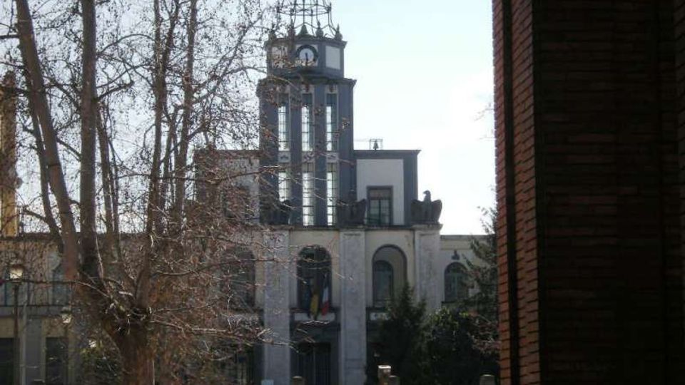 Edificio storico con facciata chiara e una torre centrale alta con finestre verticali e orologio, sormontata da una struttura metallica. In primo piano si vedono alberi spogli e una recinzione, con un muro in mattoni sul lato destro dell’inquadratura.