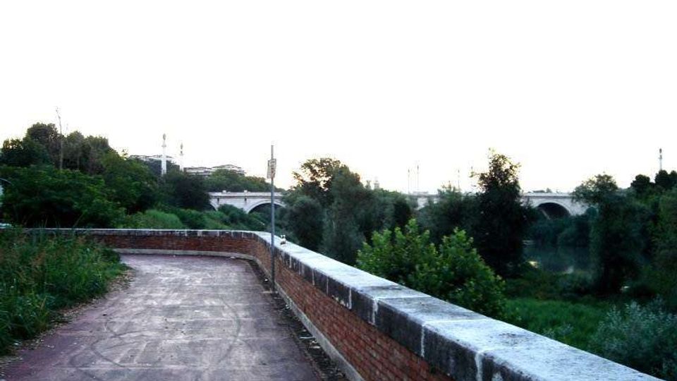 Percorso pedonale in cemento con muretto in mattoni e parapetto in pietra, affiancato da vegetazione. Sullo sfondo si vede un ponte ad archi sopra un fiume, circondato da alberi.