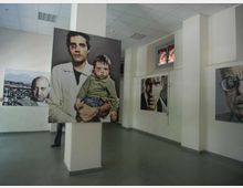Interno di una galleria o spazio espositivo con pavimento grigio e soffitto a pannelli, scandito da pilastri bianchi. Sulle pareti e su un pilastro sono appesi grandi ritratti fotografici, con una finestra in alto sul fondo.