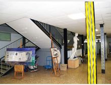 Atrio interno con scala metallica a vista, soffitto a pannelli e pavimento in graniglia, con colonne dipinte a strisce gialle e nere. Nello spazio sono presenti cavalletti con quadri e una statua su piedistallo, in un ambiente tipo scuola o centro culturale.