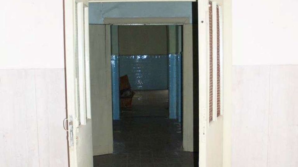 Ingresso interno con doppia porta a battente aperta che conduce a un corridoio stretto con pavimento in piastrelle scure e pareti in parte rivestite. In fondo si intravede un ambiente con piastrelle blu e luce più debole.