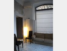 Interno di un piccolo soggiorno con divano grigio, sedie nere e lampada da terra accesa su pavimento in graniglia. Una grande finestra ad arco con inferriata decorativa e tenda a pannelli occupa la parete destra, accanto a una porta aperta su un corridoio.