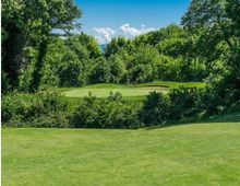 Veduta di un campo da golf con ampio prato rasato e green centrale, affiancato da un bunker di sabbia. L’area è circondata da fitta vegetazione e alberi ad alto fusto, con cielo azzurro e nuvole sullo sfondo.