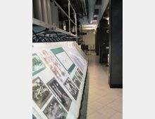 Corridoio tecnico industriale con pavimento piastrellato e grandi tubazioni e canalizzazioni a vista sul soffitto. In primo piano c’è un lungo pannello espositivo inclinato con fotografie e testi, affiancato da armadiature e macchinari scuri lungo il lato destro.