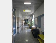 Corridoio interno di un ospedale con pavimento lucido, pareti bicolori e illuminazione a soffitto, con segnaletica sospesa che indica “Sala chirurgica”. Lungo il lato destro ci sono sedie d’attesa, piante in vaso e finestre ad arco che illuminano il passaggio.