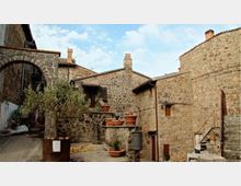 Cortile in un borgo con edifici in pietra e tetti in coppi, con scale esterne, piccoli balconi e lampioni a muro. Al centro ci sono vasi in terracotta e una pianta in una fioriera, su pavimentazione in pietra sotto un cielo parzialmente nuvoloso.