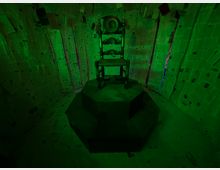 Stanza immersiva illuminata di verde, con pareti e soffitto rivestiti da fogli di carta disposti in modo irregolare. Al centro, una sedia in legno intagliato è collocata su una pedana ottagonale scura.