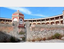 Cortile interno di una struttura fortificata con porticati ad archi su due livelli e pareti intonacate. In primo piano un basso muro in pietra e un cancello metallico, sullo sfondo una piccola torretta ottagonale rossa e bianca con cupola sotto un cielo azzurro.
