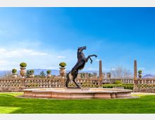 Fontana circolare in pietra con statua in bronzo di un cavallo impennato al centro di un giardino formale. Sullo sfondo si vede una balaustra in pietra con vasi ornamentali e, oltre, colline lontane sotto un cielo azzurro.