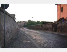 Strada urbana in pavé che curva leggermente, affiancata da alti muri in muratura e intonaco con segnaletica stradale. Sullo sfondo si intravedono edifici e alberi lungo il margine della carreggiata.