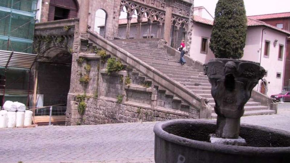 Scalinata in pietra di un edificio storico con archi decorati e portale, affacciata su una piazza lastricata. In primo piano si vede una grande fontana in pietra con un vaso centrale e una pianta potata, con impalcature sul lato sinistro dell’edificio.