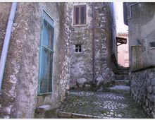 Vicolo stretto in un borgo, con pavimentazione in pietra e scalini che salgono tra edifici in muratura grezza. Sulle pareti si vedono una grande porta con grata metallica azzurra, una finestra con scuri e tubazioni a vista; a destra c’è un muretto in pietra con rete metallica.