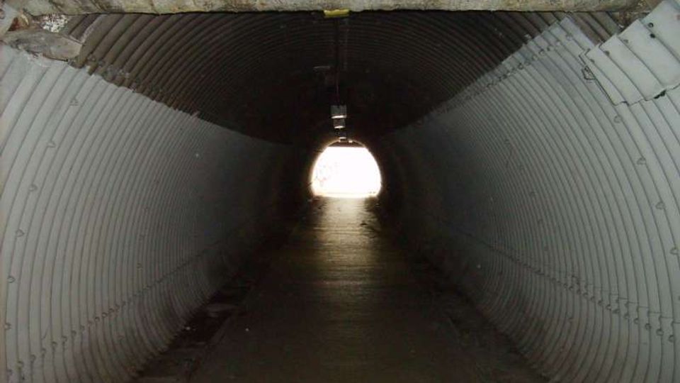 Interno di un tunnel sotterraneo con pareti e soffitto in lamiera ondulata, illuminato debolmente da una lampada centrale. In fondo si vede l’uscita circolare molto luminosa che dà sull’esterno.