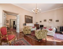 Ampio soggiorno arredato in stile classico con pavimento in parquet, tappeti persiani e salotto composto da divani e poltrone a motivi floreali. Al soffitto è appeso un lampadario in ottone e sulle pareti chiare sono presenti quadri, con una porta doppia che conduce a una sala da pranzo.