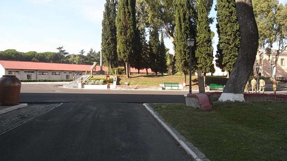 Viale asfaltato che conduce a un’area verde con alti cipressi e grandi alberi, con panchine e un lampione lungo il percorso. Sullo sfondo si vede un edificio basso con tetto rosso e una piccola scalinata in pietra.