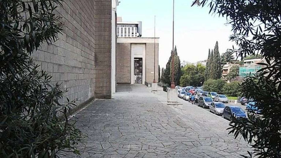 Ampio marciapiede in pietra che costeggia un grande edificio moderno rivestito in blocchi, con ingresso arretrato e alti pennoni. Sul lato destro corre una strada con auto parcheggiate, alberi e cespugli che incorniciano la vista.