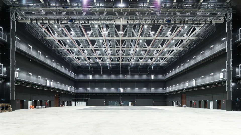 Ampio spazio interno tipo studio di produzione o palazzetto industriale, con pareti nere e passerelle perimetrali su più livelli con ringhiere metalliche. Soffitto alto con griglia di travi e tralicci illuminati, e grande pavimento libero in cemento.