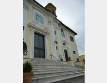 Facciata di un edificio storico in stile barocco color crema e bianco, con cornici decorative attorno a finestre e portali. Un’ampia scalinata in pietra conduce a un grande ingresso con cancello scuro, con piccoli vasi di piante ai lati e un campanile visibile sul tetto.