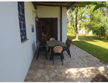 Portico di una casa con pareti bianche e tettoia, pavimentato in pietra, con tavolo rettangolare e sedie da esterno vicino a una porta in legno aperta. A destra si vede un giardino erboso con alberi e vegetazione, illuminato dalla luce del sole.
