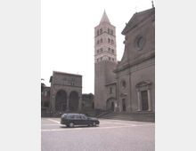 Piazza lastricata con una chiesa in pietra e ampia scalinata d’ingresso, affiancata da un alto campanile a fasce orizzontali e da un edificio con portico ad archi. Sullo sfondo si vedono altri edifici storici che delimitano lo spazio urbano.