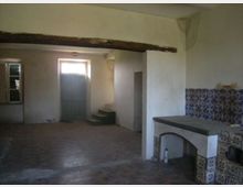 Interno di una vecchia cucina rurale con pavimento in mattoni, travi a vista e pareti intonacate. A destra c’è un grande banco in muratura rivestito di piastrelle decorative, mentre sul fondo si vedono una porta, una finestra e un piccolo gradino che conduce a un vano.