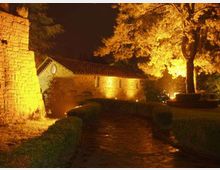 Percorso in pietra in un giardino con siepi basse, illuminato da luci notturne che evidenziano un edificio in pietra con tetto in tegole e una torre muraria a sinistra. Un grande albero sulla destra è illuminato dal basso, con sfondo di cielo scuro.