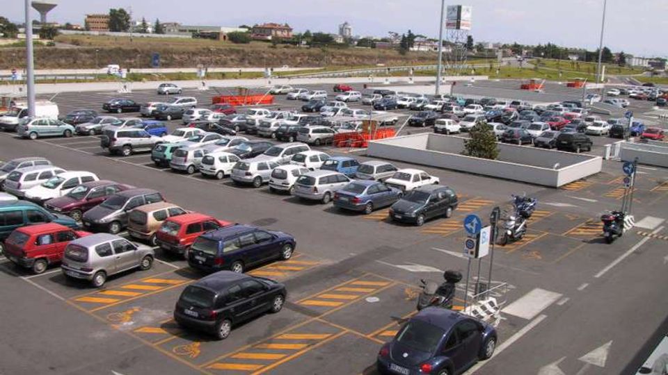 Ampio parcheggio asfaltato all’aperto con molte auto parcheggiate in file ordinate, segnaletica orizzontale con frecce direzionali e stalli riservati. Sullo sfondo si vedono strade di scorrimento, lampioni alti e un’area collinare con edifici sparsi.