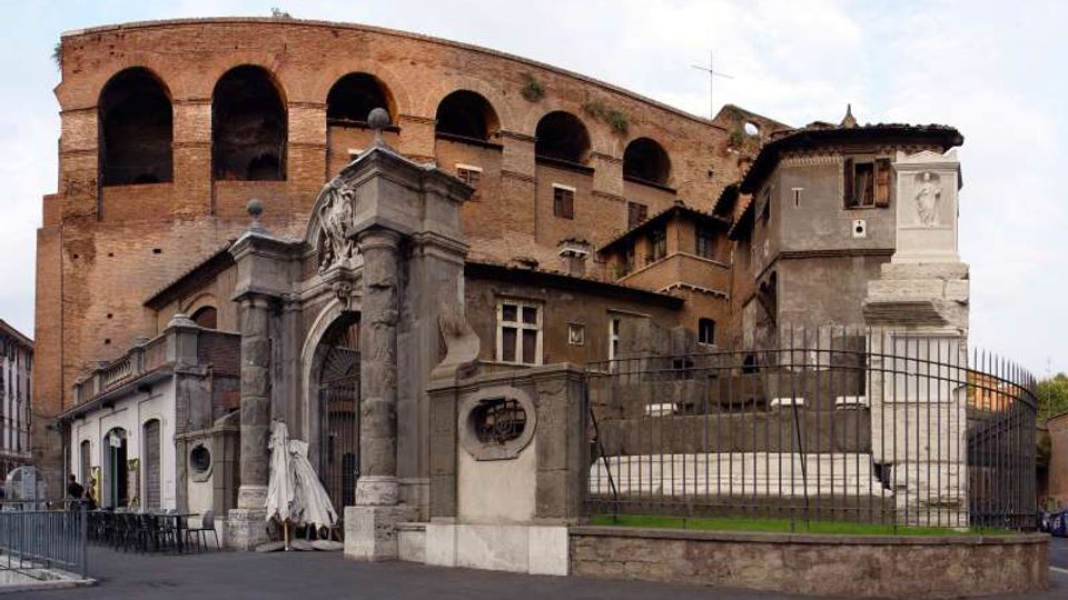 Veduta esterna di un grande edificio storico in mattoni con arcate sovrapposte, affiancato da strutture più basse e da un ingresso monumentale in pietra con portale ad arco. In primo piano si vede una recinzione metallica lungo un marciapiede e un’area rialzata con prato.