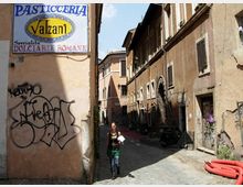 Vicolo stretto con pavimentazione in ciottoli tra edifici in muratura color ocra con persiane, finestre e segni di usura. In primo piano c’è un grande cartello “Pasticceria Valzani” su una parete con graffiti, mentre lungo la strada si vedono un portone ad arco e alcuni oggetti appoggiati ai muri.
