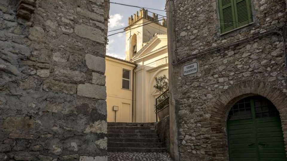 Vicolo in pietra con scalinata e pavimentazione in ciottoli tra edifici in muratura, con arco e portone verde e finestra con persiane. In fondo si intravede la facciata chiara di una chiesa con campanile e cavi sospesi sopra la strada.