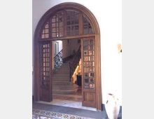 Grande portale ad arco in legno con ante a vetri che si apre su una scala interna in pietra con ringhiera in ferro battuto, in un ambiente dai muri chiari e pavimento piastrellato decorato.