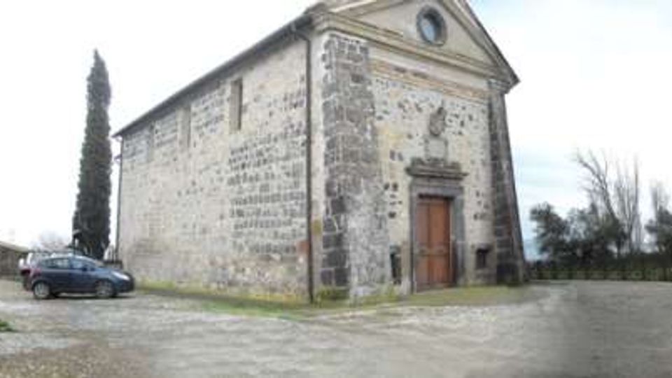 Piccola chiesa in pietra con facciata a timpano e portale centrale, situata su uno spiazzo di ghiaia. Intorno si vedono cipressi e altri alberi, con una strada e un muretto lungo il bordo dell’area.