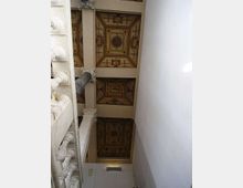 Soffitto interno decorato con riquadri a cassettoni dipinti con motivi geometrici e medaglioni centrali, sostenuti da travi e colonne. A sinistra si vede una balaustra in stile classico, mentre a destra una parete bianca corre lungo un vano stretto verso l’alto.