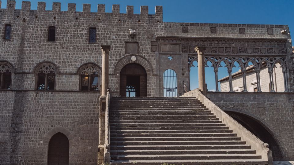 Facciata in pietra di un palazzo medievale fortificato con merli, finestre ad arco e una grande scalinata centrale che conduce a un portale. A destra si vede una loggia con arcate gotiche sorrette da colonne, sullo sfondo del cielo azzurro.
