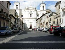 Piazza lastricata in un centro storico, con auto parcheggiate ai lati e palazzi residenziali che fiancheggiano la strada. Sullo sfondo si vede una grande chiesa barocca bianca con facciata simmetrica, portale centrale e due campanili laterali.