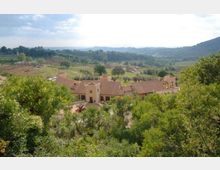 Vista panoramica di un complesso di edifici rurali con tetti in tegole e piccoli campanili, circondato da vegetazione. Sullo sfondo si estendono campi coltivati, filari di alberi e colline verdi sotto un cielo parzialmente nuvoloso.