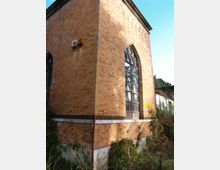 Angolo di un edificio in mattoni a vista con grande finestra ad arco e vetrate, incorniciata da decorazioni nella parte superiore. Alla base si vedono vegetazione e arbusti, con cielo sereno e altri fabbricati sullo sfondo.