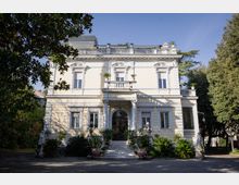 Facciata di una villa storica a due piani intonacata chiara, con ingresso ad arco e colonne, balcone centrale e ringhiere decorative sul tetto. Davanti, una scalinata con vasi e piante ornamentali conduce all’entrata, circondata da alberi e giardino.