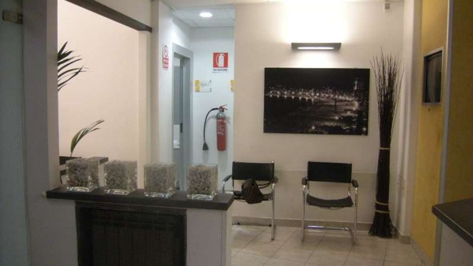 Piccola sala d’attesa interna con pavimento piastrellato e soffitto a pannelli, due sedie nere contro la parete e un quadro fotografico sopra. A sinistra si vede un bancone/reception con contenitori in vetro, mentre sul fondo ci sono una porta, un estintore a parete e un vaso alto con rami decorativi.
